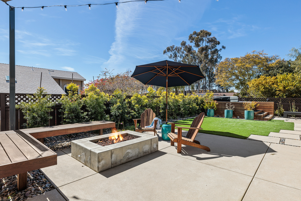 Backyard with a concrete fire pit, wood benches, Adirondack chairs and a navy umbrella