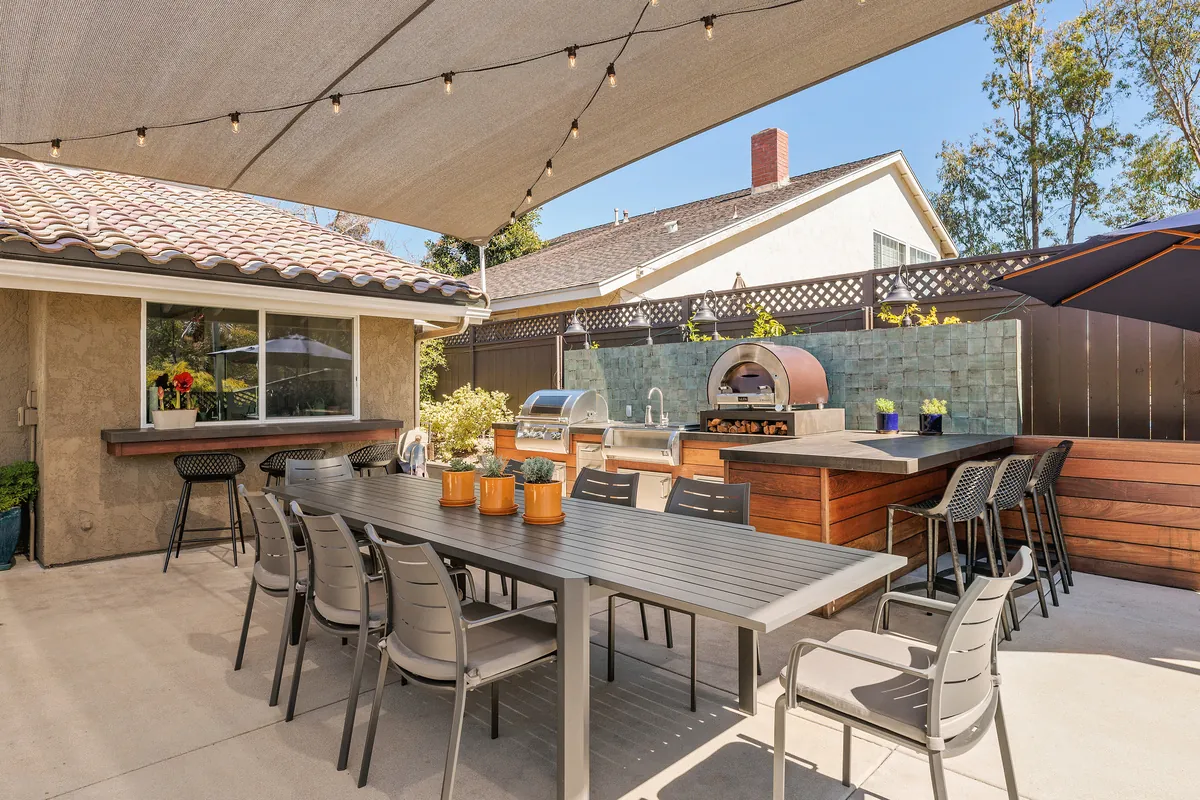 The backyard after: a sail shade with string lights, an outdoor kitchen with a pizza oven and a long dining table